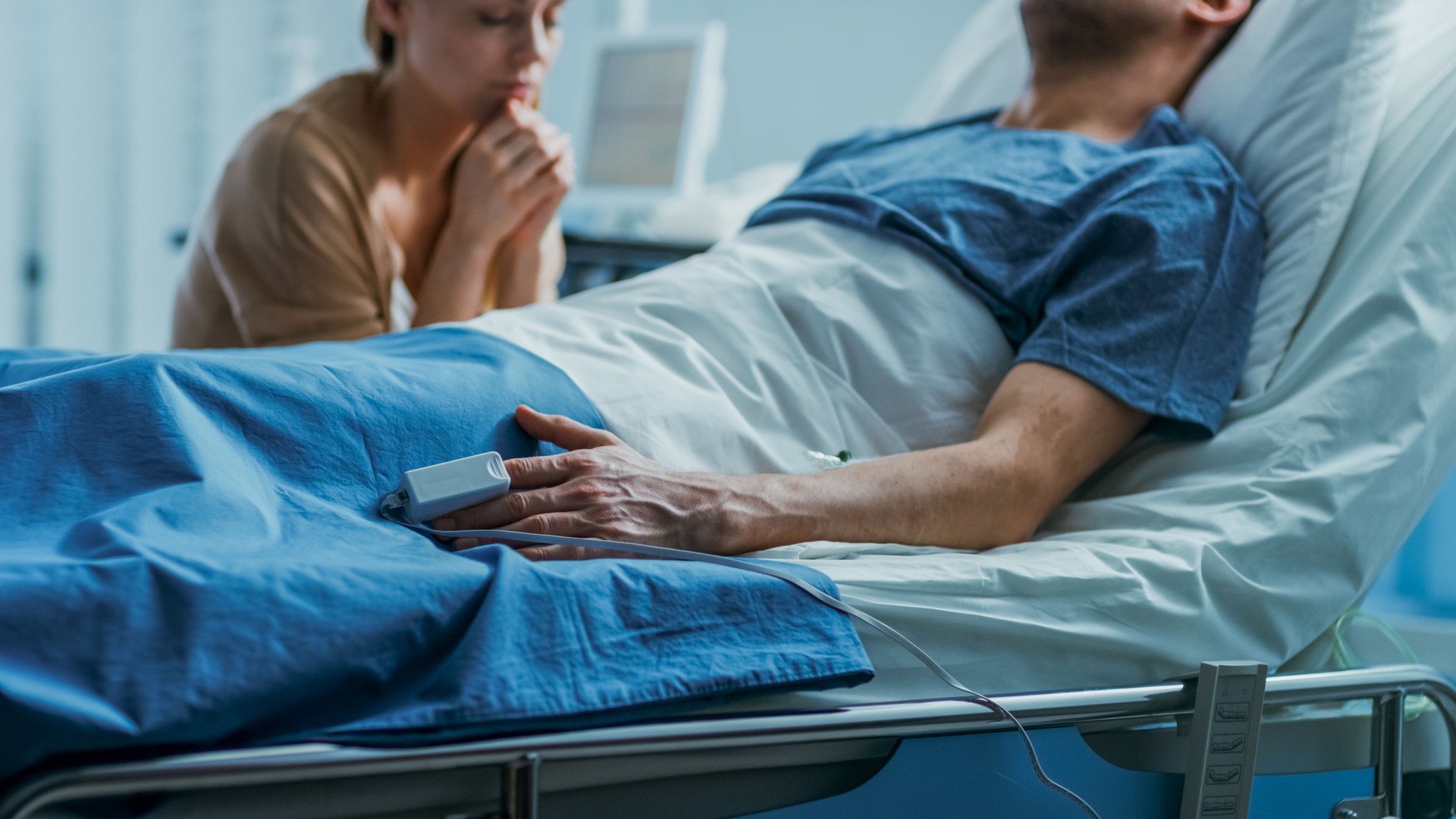 In the Hospital Sick Man Lying on the Bed, His Visiting Wife Hopefully Sits Beside Him and Prays for His Rapid Recovery. Tragic, Somber and Melancholy Scene.